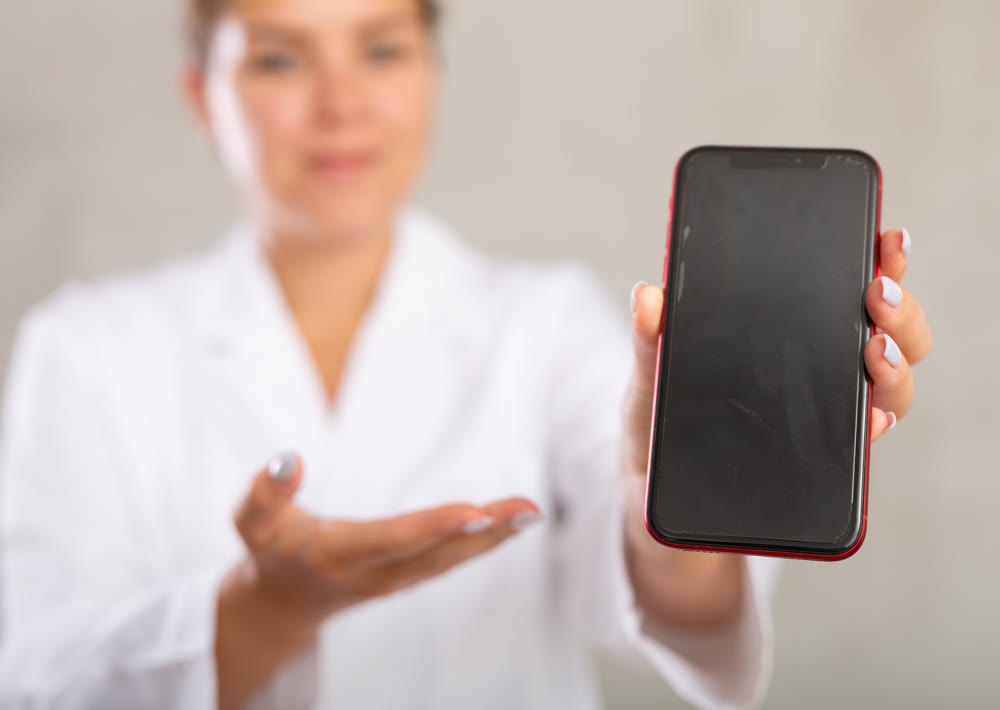 Female doctor in coat extending hand with phone and showing screen, selective focus on gadget. Online consultation concept