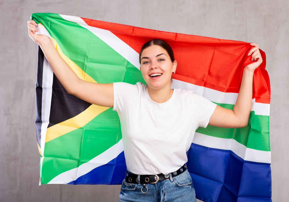 Joyous young woman holding South Africa flag against unicoloured background