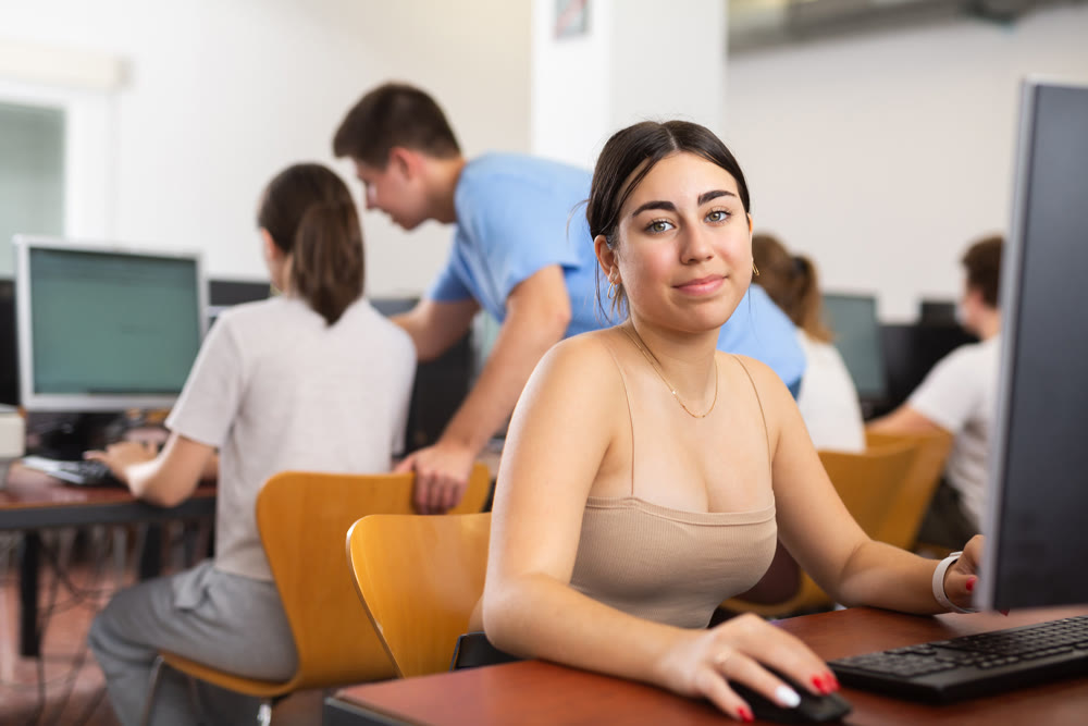 Smiling young woman sitting in computer room at workplace and looking at camera