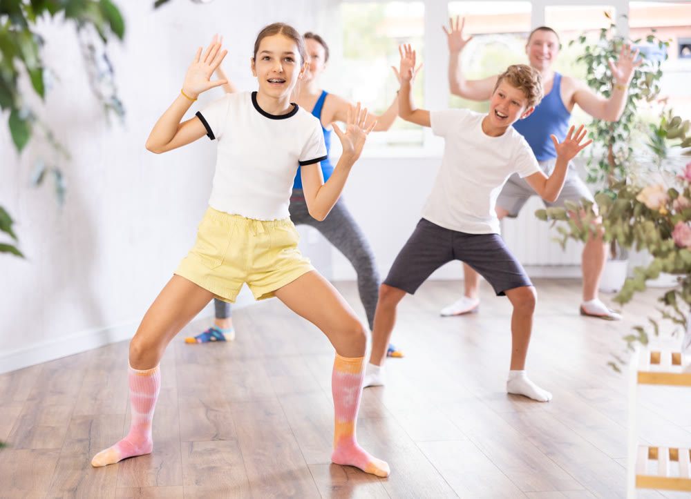 Joyful sister and brother practicing expressive dance movements against backdrop of mom and dad training together in wellness center