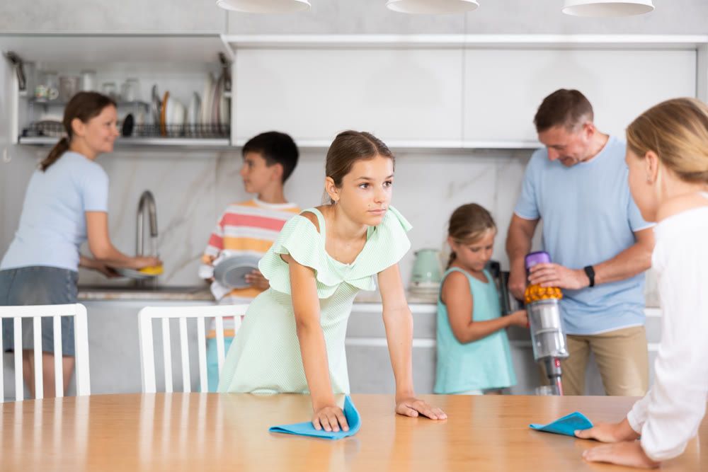 Large family is cleaning room in house - two daughters are dusting table, dad and son are vacuuming room