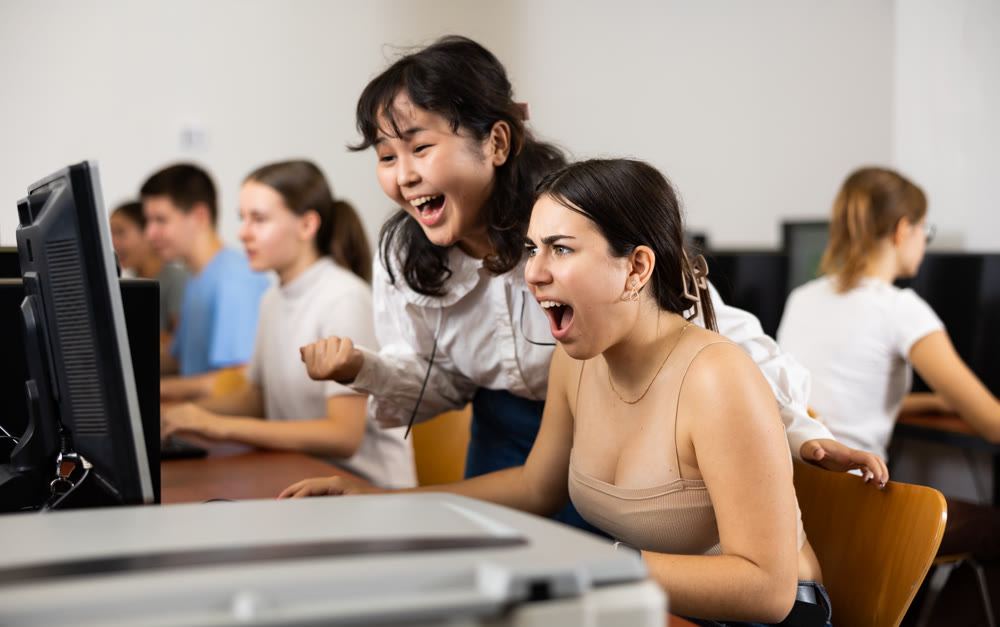Excited female classmates laughing and looking at screen of PC while learning computer science in the classroom