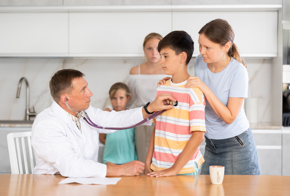 Doctor listens with phonendoscope to boy heart rate at home. Worried family in background