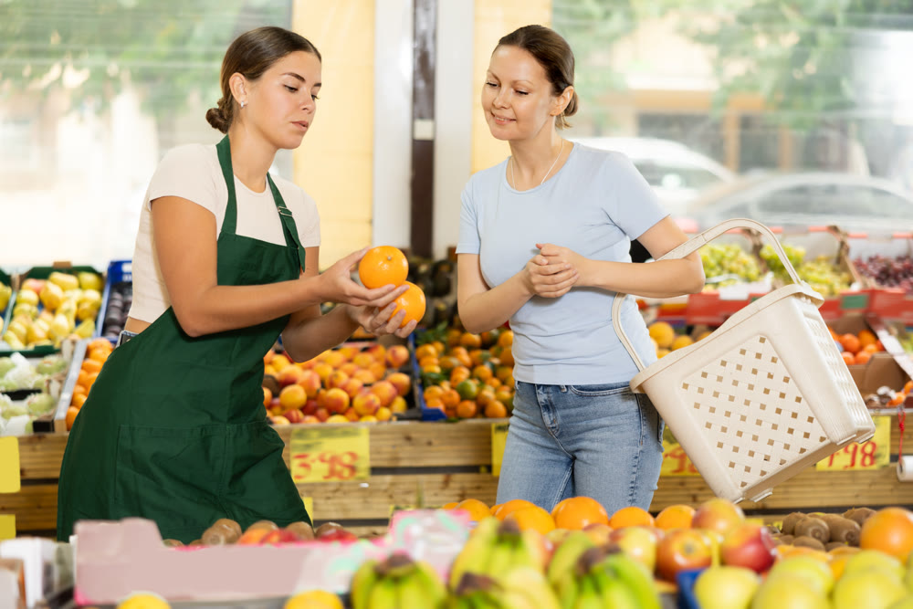 Polite salesgirl offering sweet oranges to female customer in greengrocery store