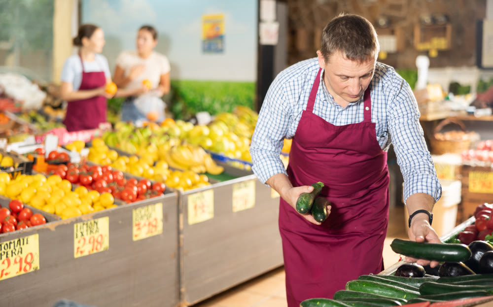 Man seller working in supermarket and lays out cucumber on counter