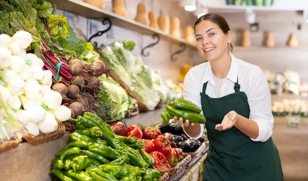 Young female owner of fruit and vegetable shop offering peppers