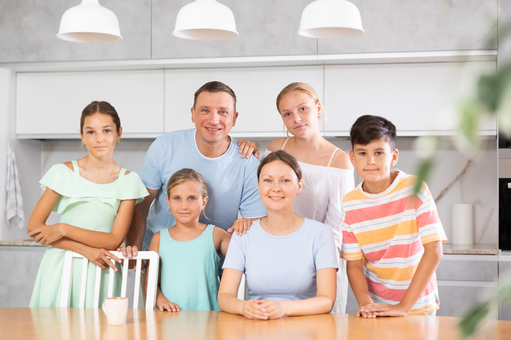 Happy dad and mum with three daughters and son is standing near kitchen table.