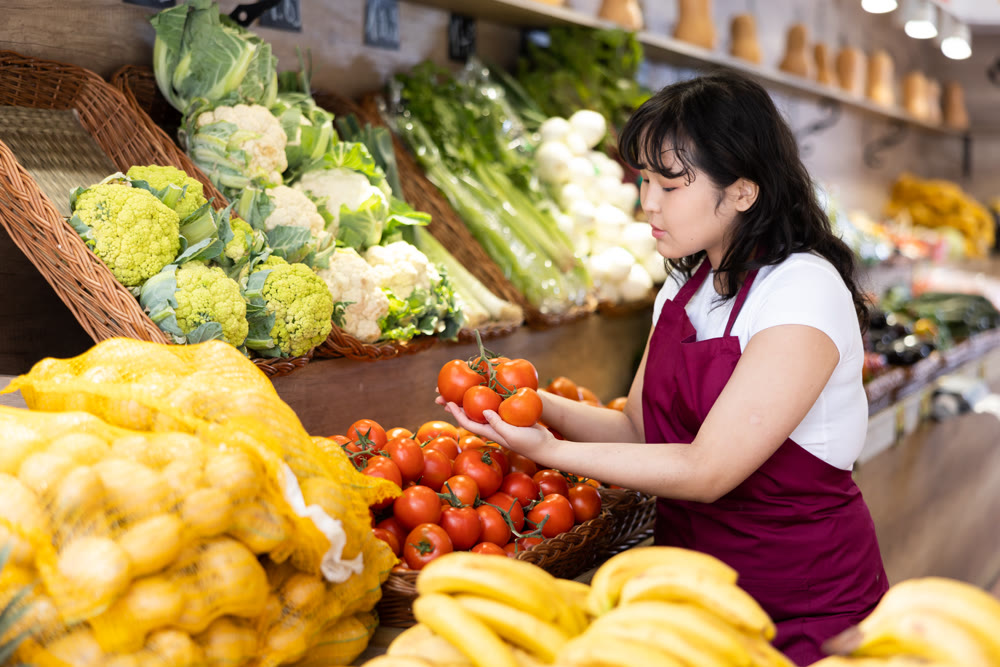 Attentive young saleswoman placing tomatoes on food stall in grocery store
