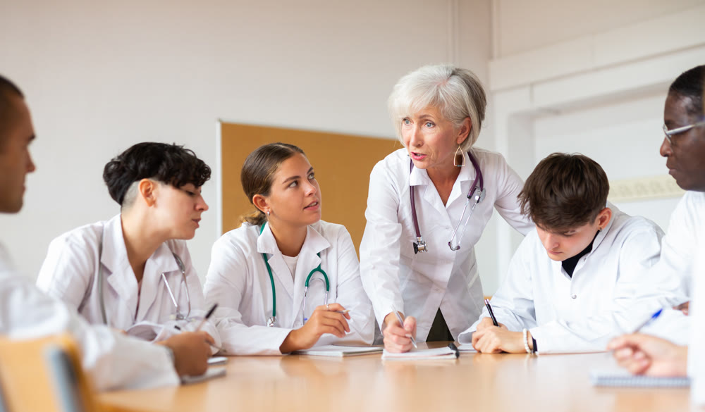 Elderly female doctor conducts training for interns of different ages at round table in auditorium of medical university