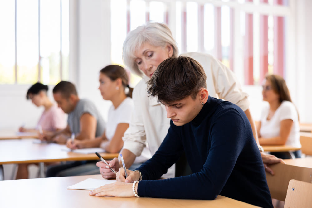 Aged female teacher helping guy student solving problem at lesson