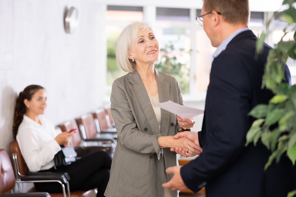 Senior woman makes deal, shaking hands with middle-aged man colleague while standing in office