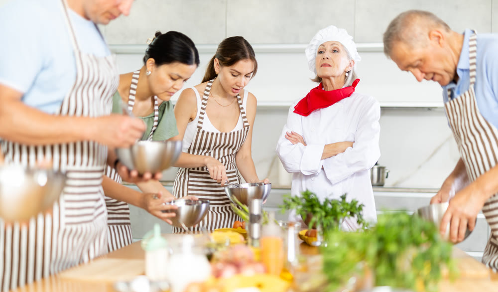 Smiling old woman chef of cooking course teaching attendees how to mix sauce