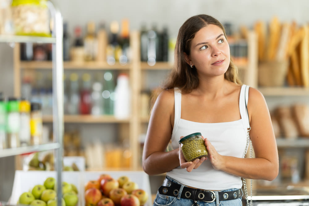 Young female customer carefully choosing can of green peas in grocery supermarket