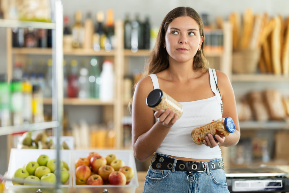 Young female shopper carefully choosing jar of canned beans in grocery supermarket