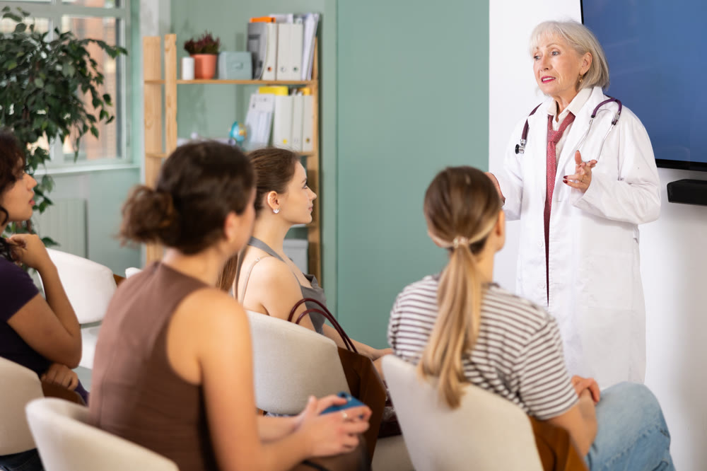 Concentrated mature female medical doctor dressed in white medical coat with stethoscope standing and talking in auditorium where sitting medical student