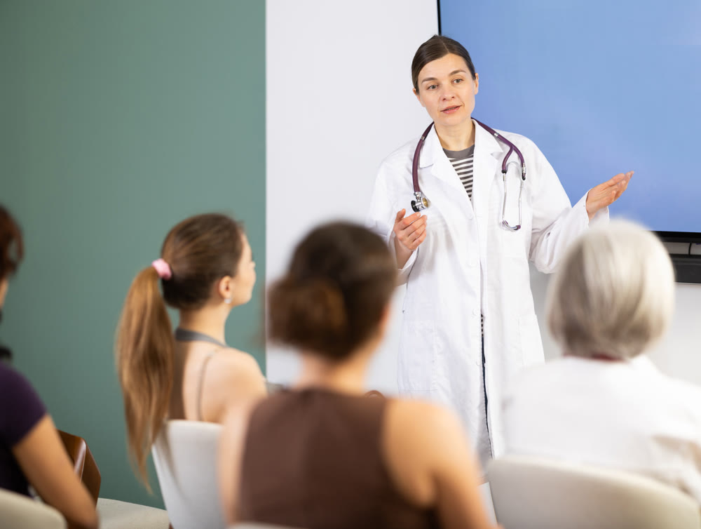 Positive young girl medical doctor dressed in white medical coat with stethoscope standing and talking in auditorium where sitting medical student